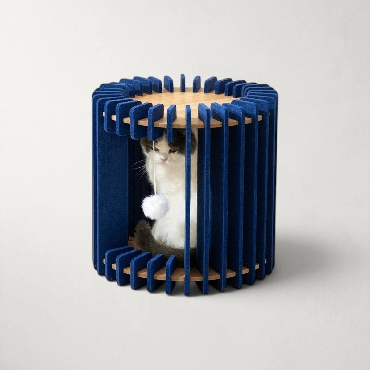 Niche pour chat Bofélin cylindrique en feutre bleu nuit et bois clair avec pompon suspendu et chat à l'intérieur