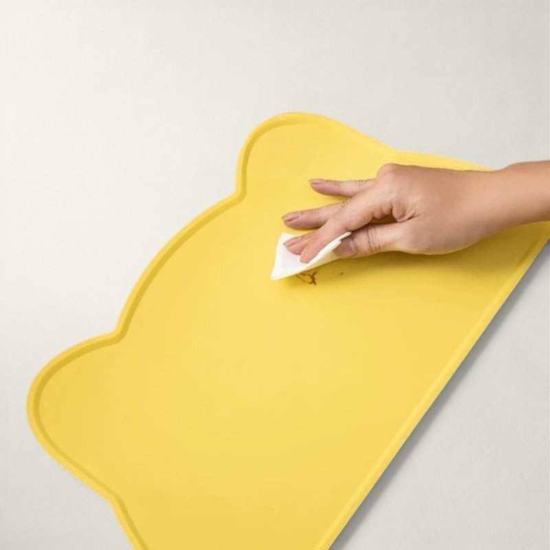 Tapis de gamelle Bofélin en silicone jaune en forme de tête de chat, nettoyé avec une éponge pour protéger le sol.